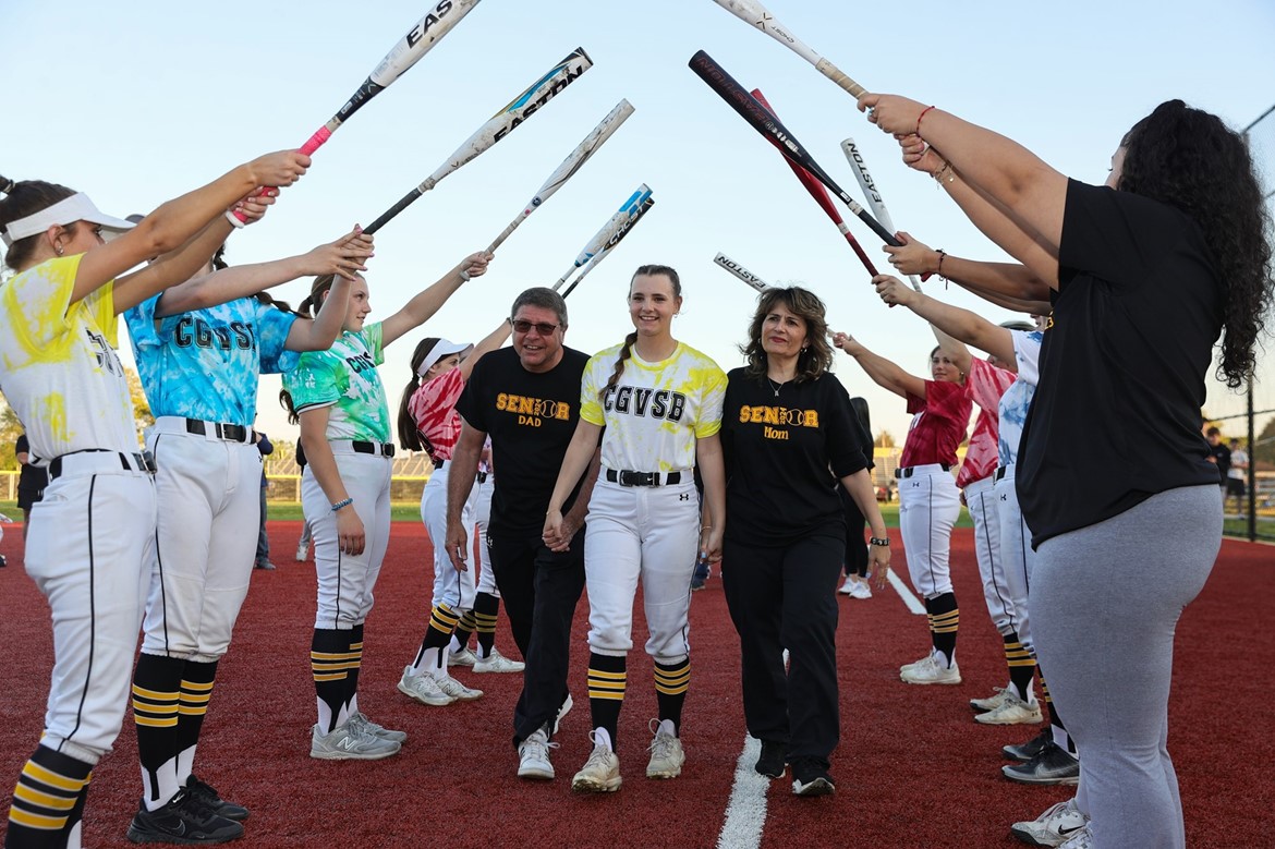 Softball senior and parents walk through canopy of teammates holding bats during postgame ceremony.